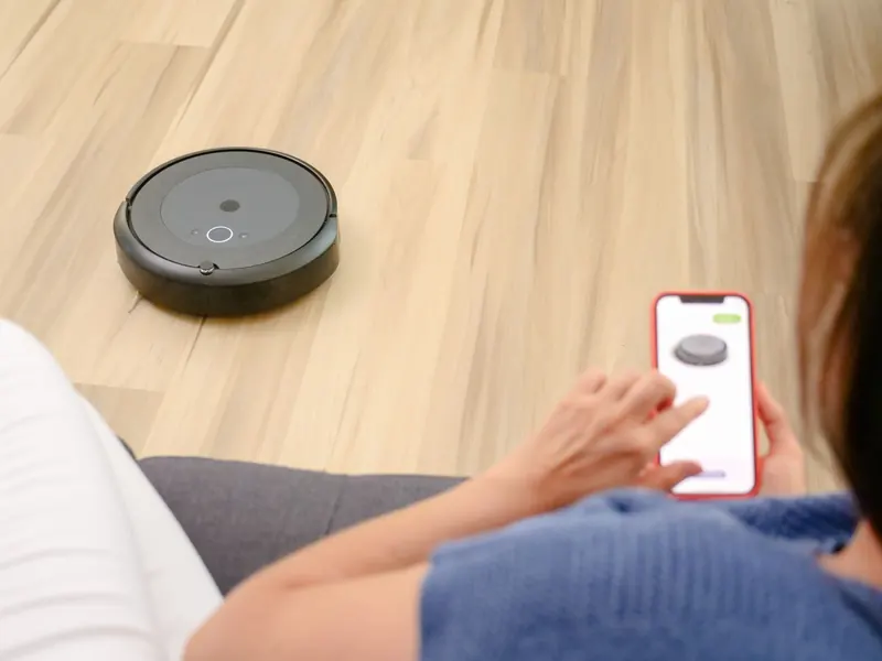 A person uses a smartphone app to control a robotic vacuum on a hardwood floor, showcasing smart home technology
