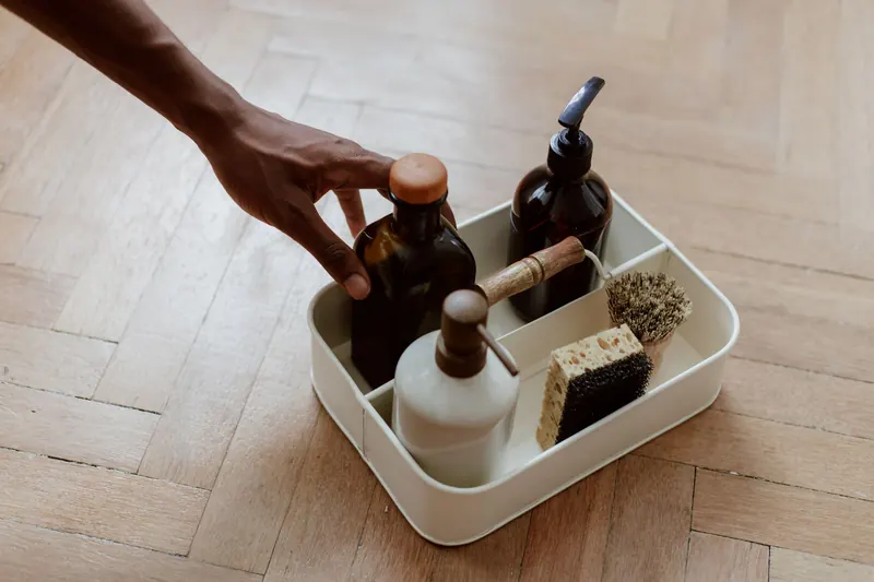 A hand reaching for a cleaning set with bottles and brushes on a wooden floor