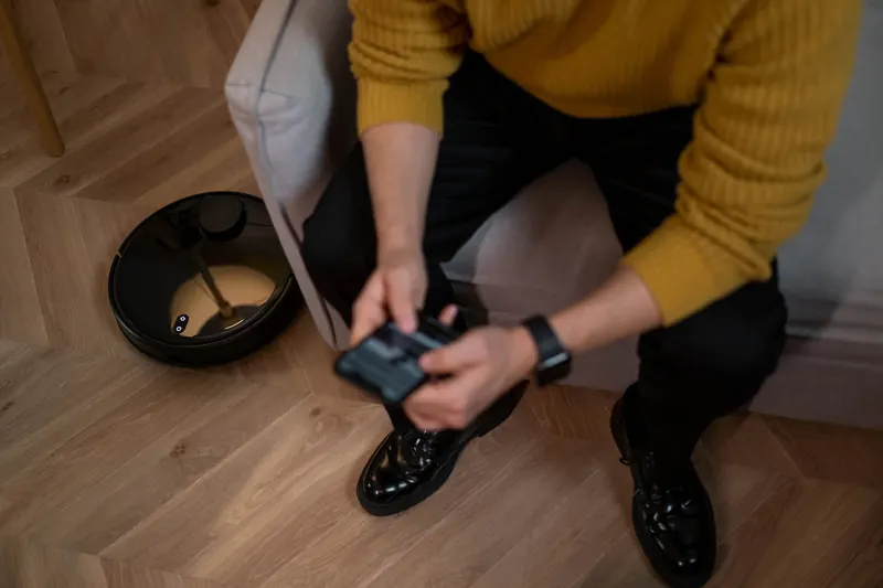 Man in yellow sweater using remote to control robotic vacuum on wooden floor
