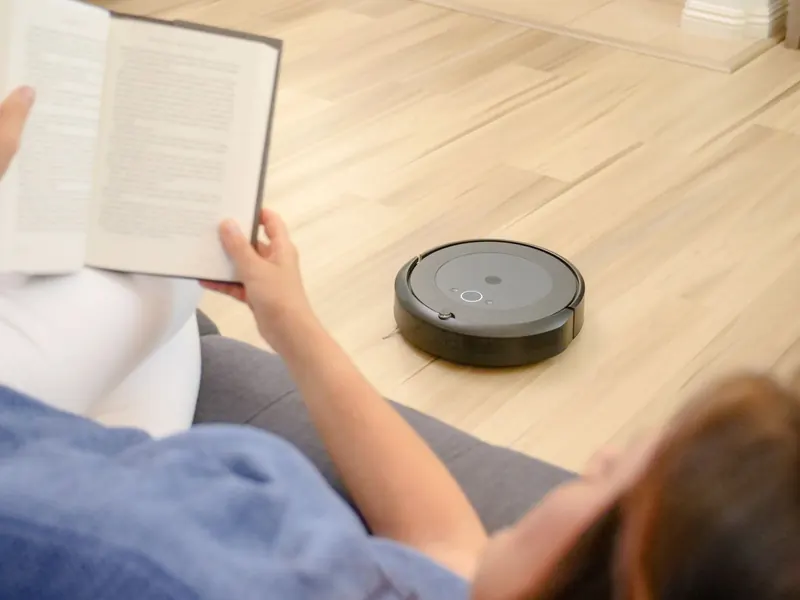 A woman relaxes on a sofa reading while a robotic vacuum navigates the floor