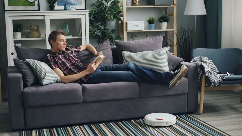 A man reading a book on a sofa, while a robotic vacuum cleans the living room floor
